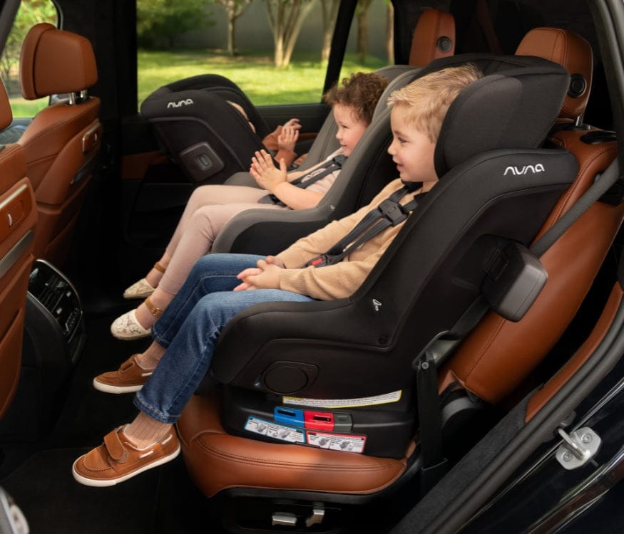 Two children sit in black and gray Nuna car seats in a car with brown leather interior. They are wearing seatbelts and looking forward.