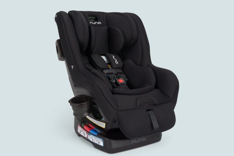 A black Nuna car seat stands angled slightly left on a blue-gray background. The seat is equipped with a cup holder and adjustable straps.