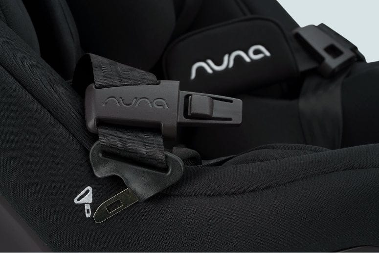 Close-up of a black Nuna car seat. The seatbelt is buckled, with the Nuna logo visible on the buckle and seat pad.