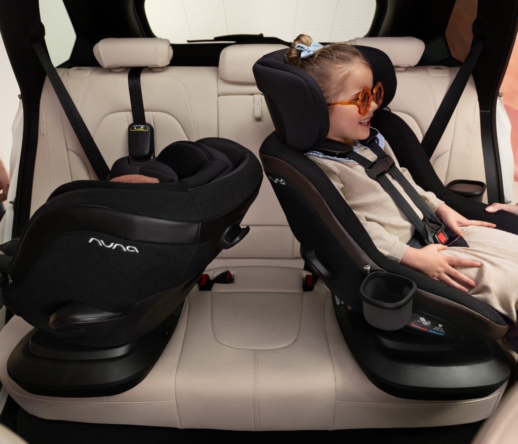 Girl smiling in black Nuna car seat, wearing orange sunglasses, beside empty matching seat in beige car interior.