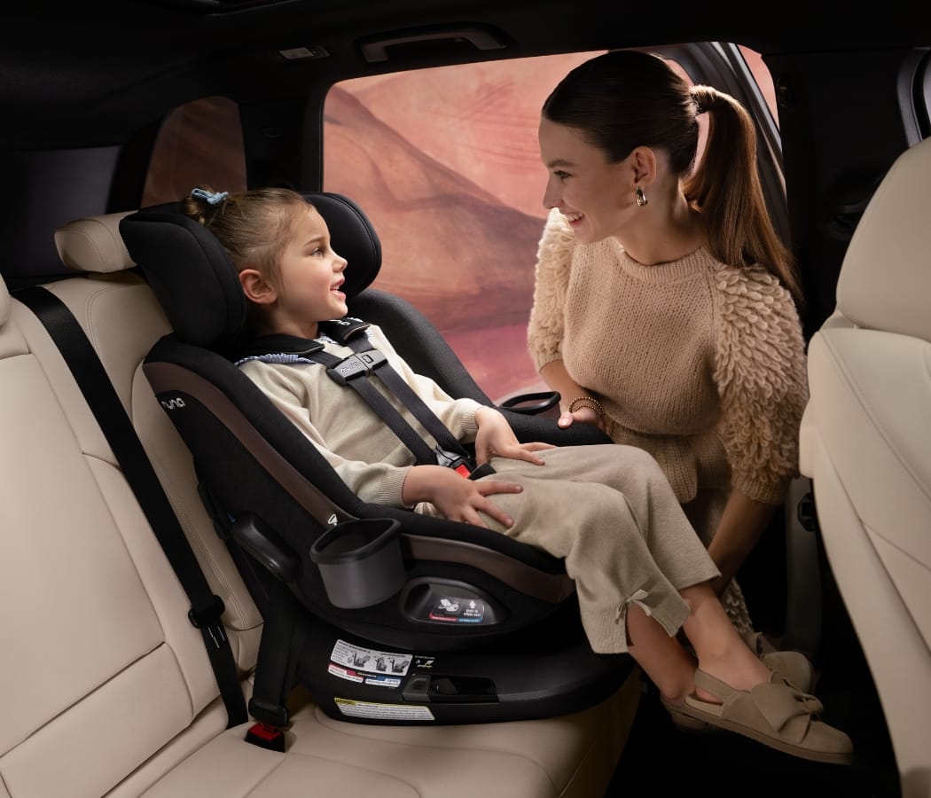 Mother smiles at child in black car seat inside beige interior, warm lighting, desert view outside.