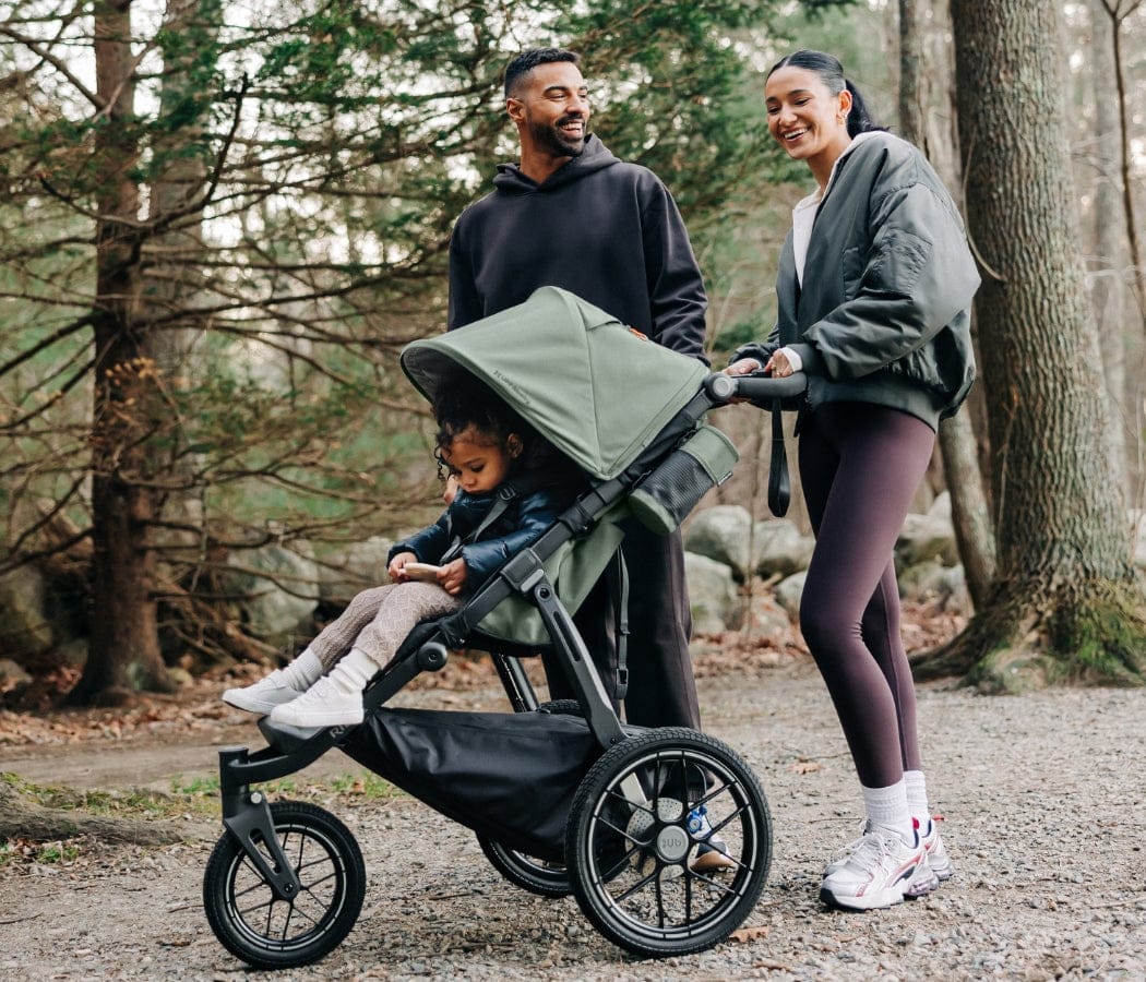 Family of three on forest path, mother pushing green stroller with child, father walking beside, all smiling.