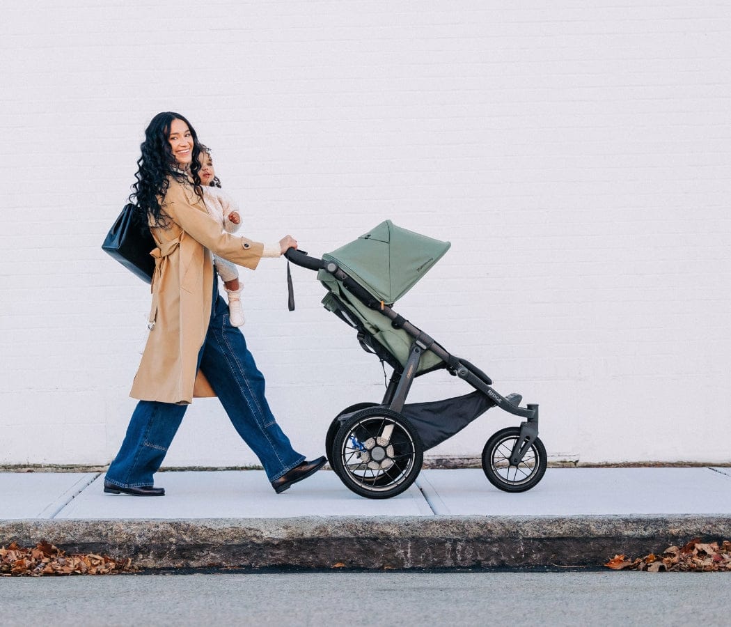 Woman in tan coat pushes green stroller while carrying child, walking on sidewalk beside white wall.