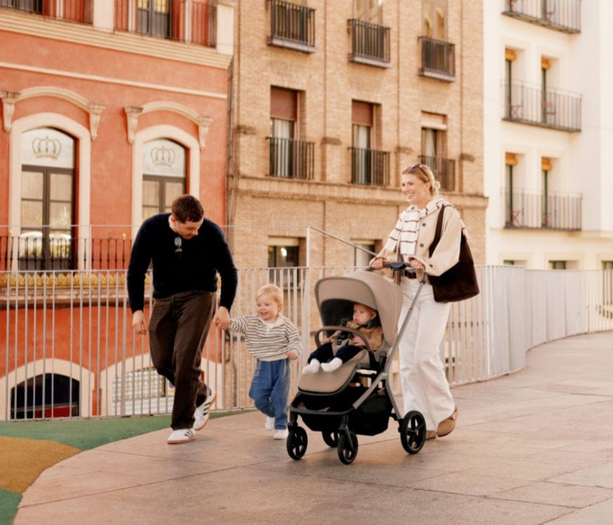 A family walks on a city street: a mother pushes a stroller with a baby, a father holds a toddler's hand. Brick buildings rise in the background.