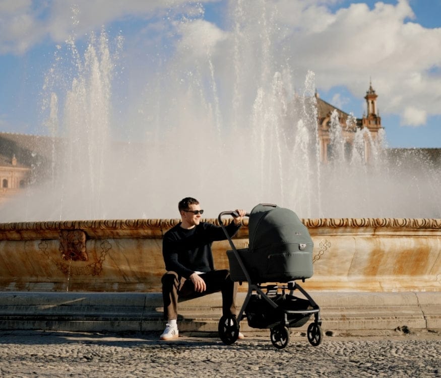 A man sits by a large fountain with a stroller, the sun shining behind him on a tan building.