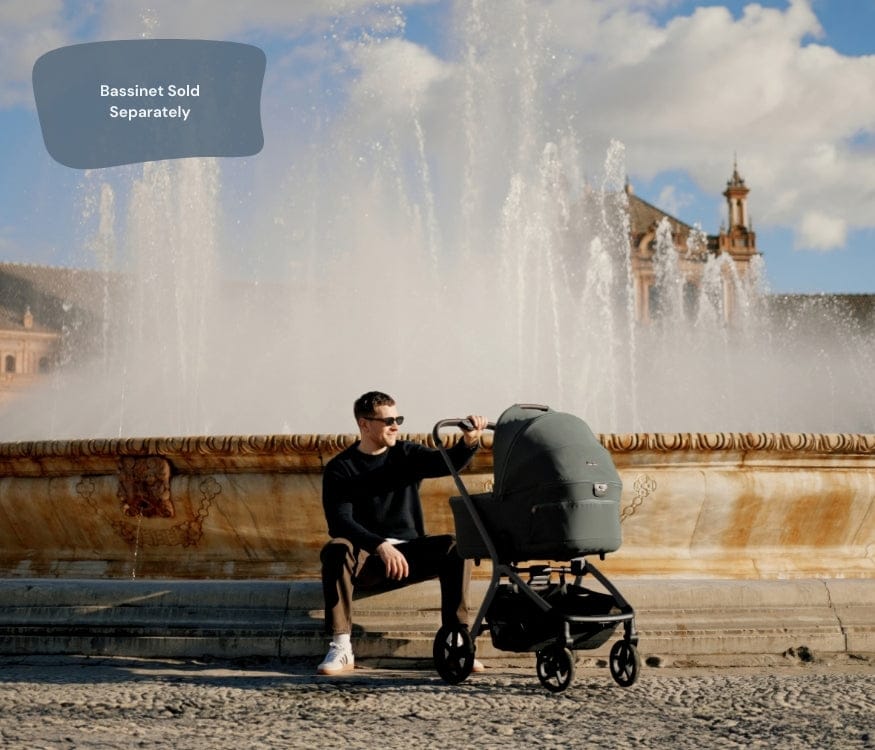 A man sits with a stroller beside a fountain, with a building visible in the background. The text "Bassinett Sold Separately" appears in the upper left.
