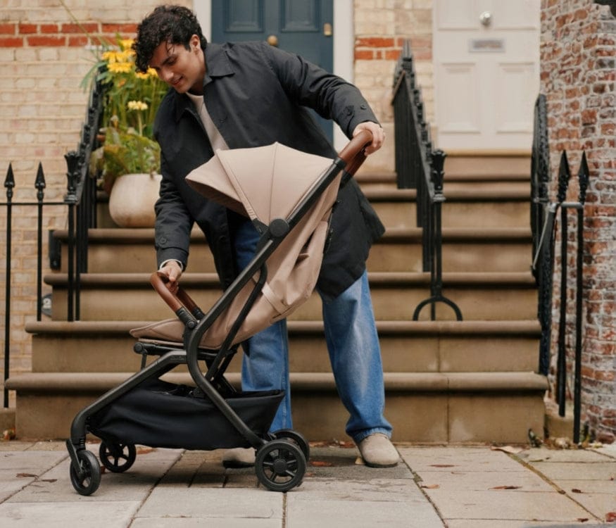 A man in a trench coat folds a beige stroller on a sidewalk with steps and potted flowers in the background.