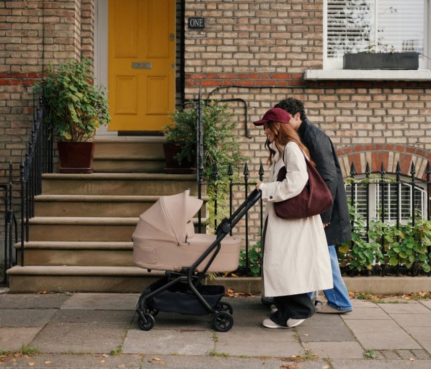 A couple strolls past a brick house, pushing a beige stroller along a sidewalk. The house has a yellow door and potted plants on the steps.
