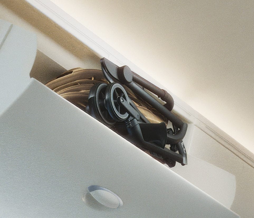 A folded black stroller and tan fabric sit inside an overhead compartment, viewed from a low angle. The compartment is light gray.
