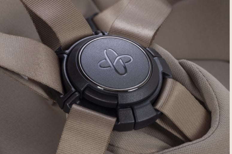 Close-up of a baby car seat harness with a black central lock, brown straps, and a tan fabric background.