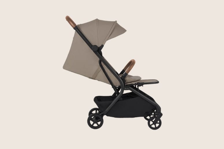 A beige and black stroller sits on a beige surface, viewed from the side. The stroller has a black frame, a beige canopy, and brown accents.