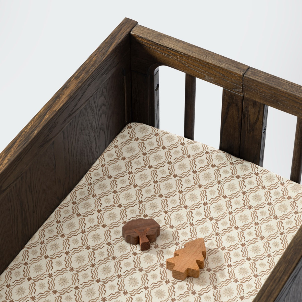 Babyletto dark wood crib with patterned cream and brown fitted sheet and two wooden tree toys