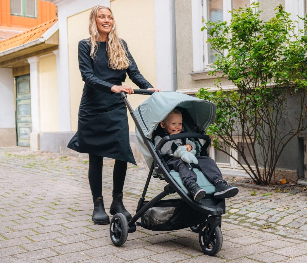Woman in black dress pushes stroller with smiling baby on cobblestone street, green canopy, urban background.
