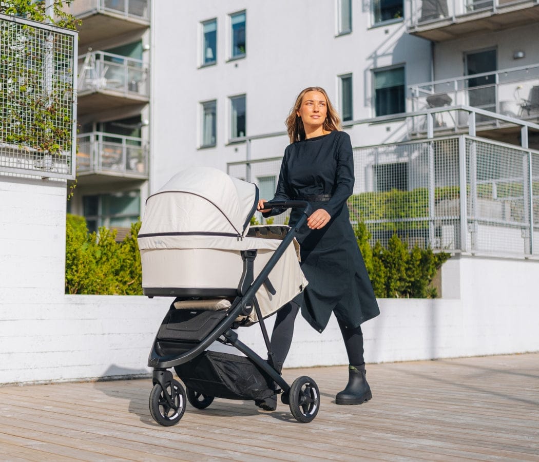 Woman in black dress pushes beige baby stroller on wooden deck, modern white building in background.
