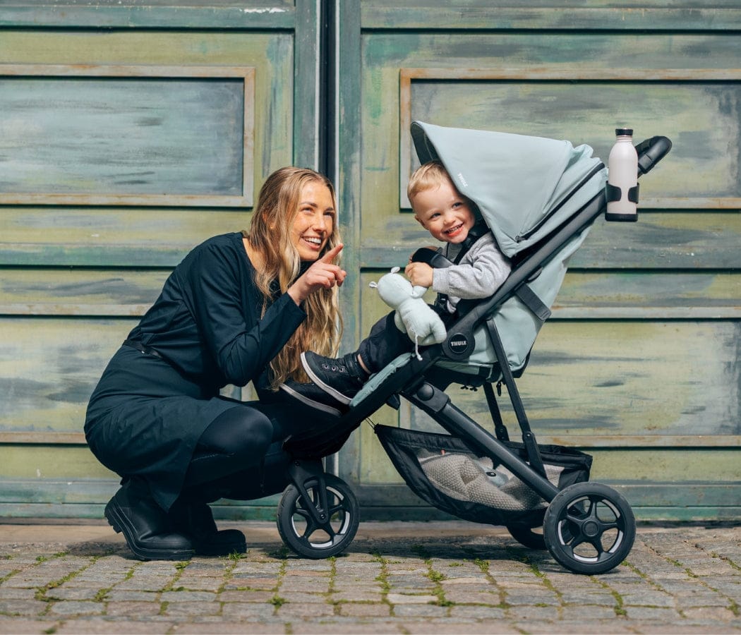 Woman crouching beside smiling toddler in light blue stroller, both pointing, against weathered green wooden wall.