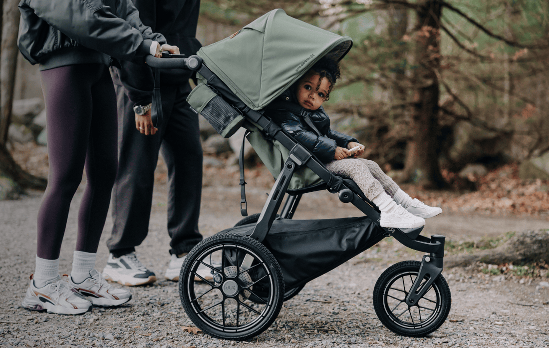 child in uppababy ridge v2 stroller in green in a forrest setting with parents pushing stroller.