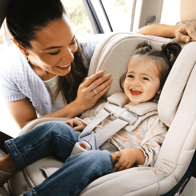 A smiling toddler is buckled in a car seat, with a woman looking down and smiling at them. Natural lighting.
