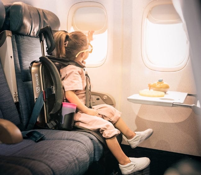 A young girl on an airplane, looking out the window, with a backpack behind her and food on the tray table. Interior, bright daylight. "WAYB" is visible on the backpack.
