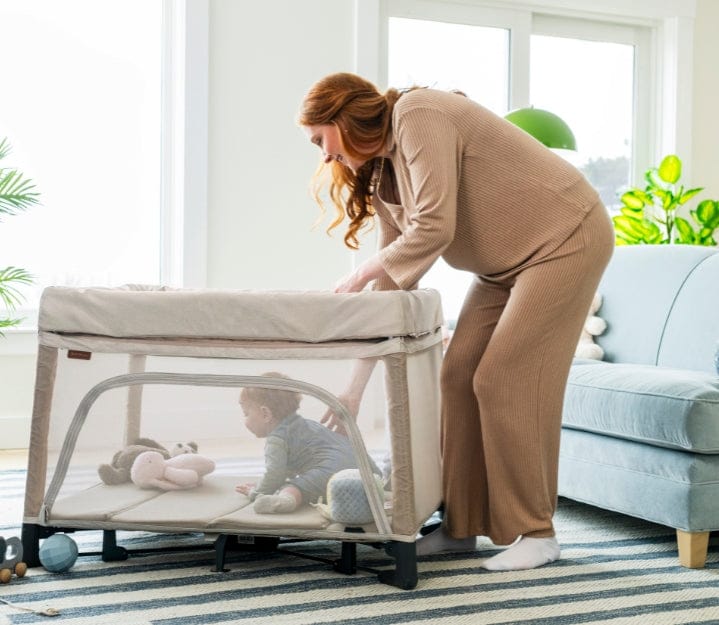 A pregnant woman leans over a playpen containing a baby and several toys in a brightly lit room.