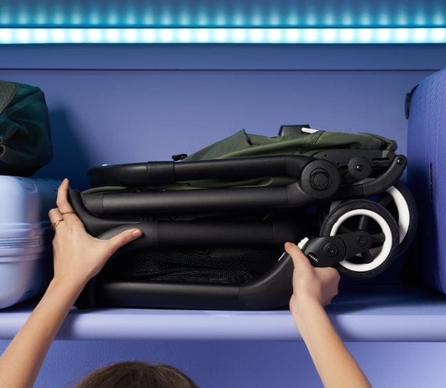 A folded green and black stroller sits on a purple shelf between two pieces of luggage, being held by two hands.