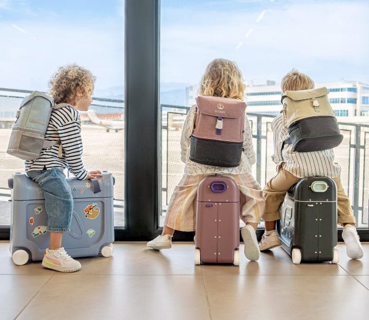 Three children sit on wheeled suitcases, gazing out a large airport window with backpacks. They appear hopeful and excited.