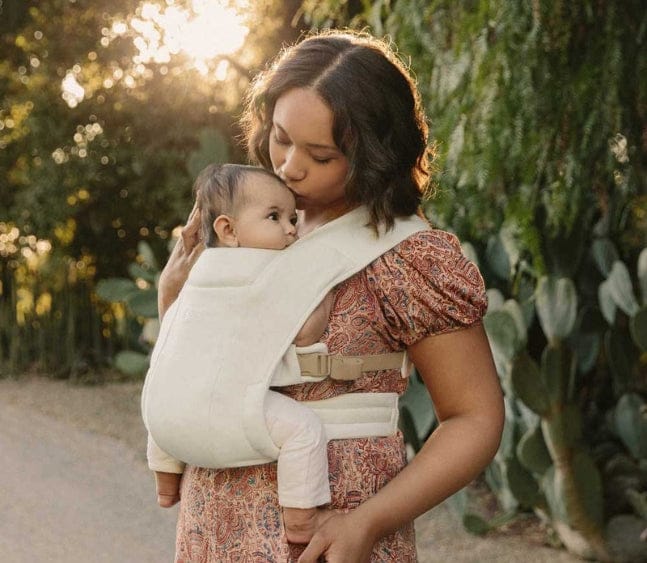 A woman with dark hair holds a baby in a carrier, kissing its forehead. The scene is warmly lit, with trees in the background.