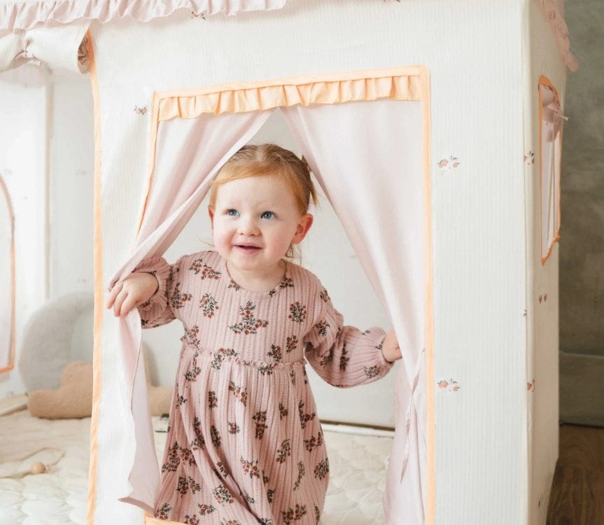 A young, fair-skinned child with red hair smiles while standing in the opening of a white and pink playhouse. The child is wearing a mauve floral dress.