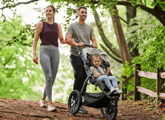 Family walking in forest, man pushing stroller with child, woman beside, green trees, natural light, peaceful outdoor scene.