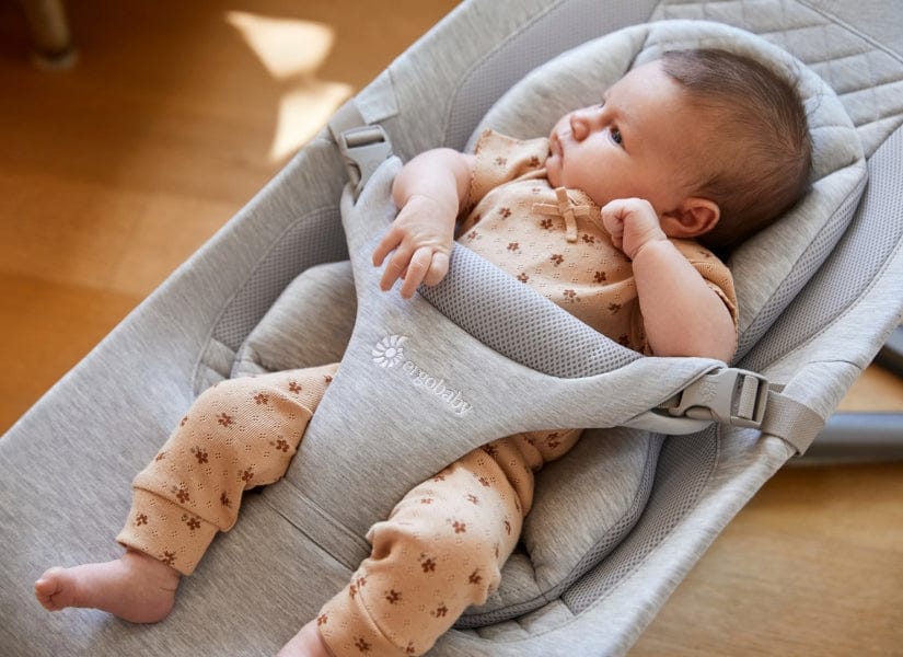 Baby in tan floral onesie resting in gray Ergobaby carrier, lying on wooden floor, sunlight streaming in.