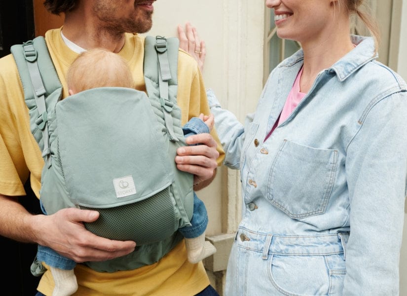 Man in yellow shirt carries baby in green Ergobaby carrier, woman in denim overalls smiles beside him, warm outdoor lighting.