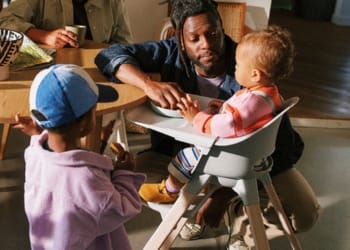 Father feeding toddler in a high chair while another child stands nearby