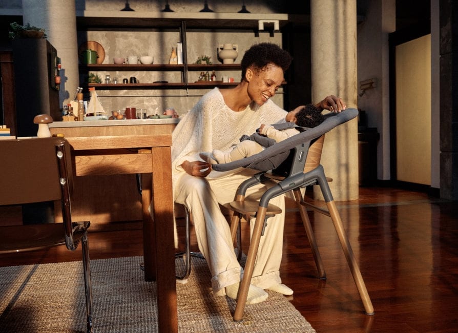 Woman in white sweater sits at wooden table, smiling as she rocks baby in gray high chair with wooden legs; warm sunlight streams through window, illuminating rustic kitchen shelves in background.