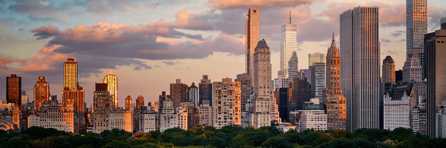 City skyline at sunset with tall skyscrapers and green trees in foreground, warm golden light and pink clouds.