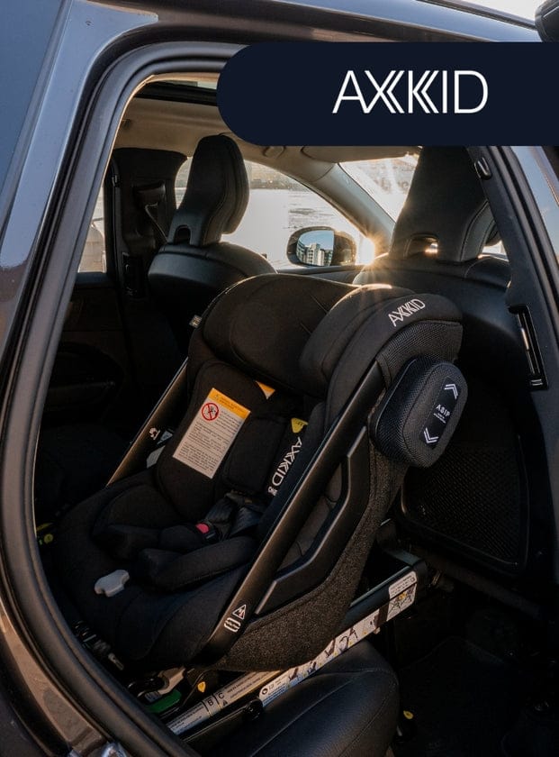 Black AXKID child car seat installed in vehicle backseat, viewed through open door with sun flare.