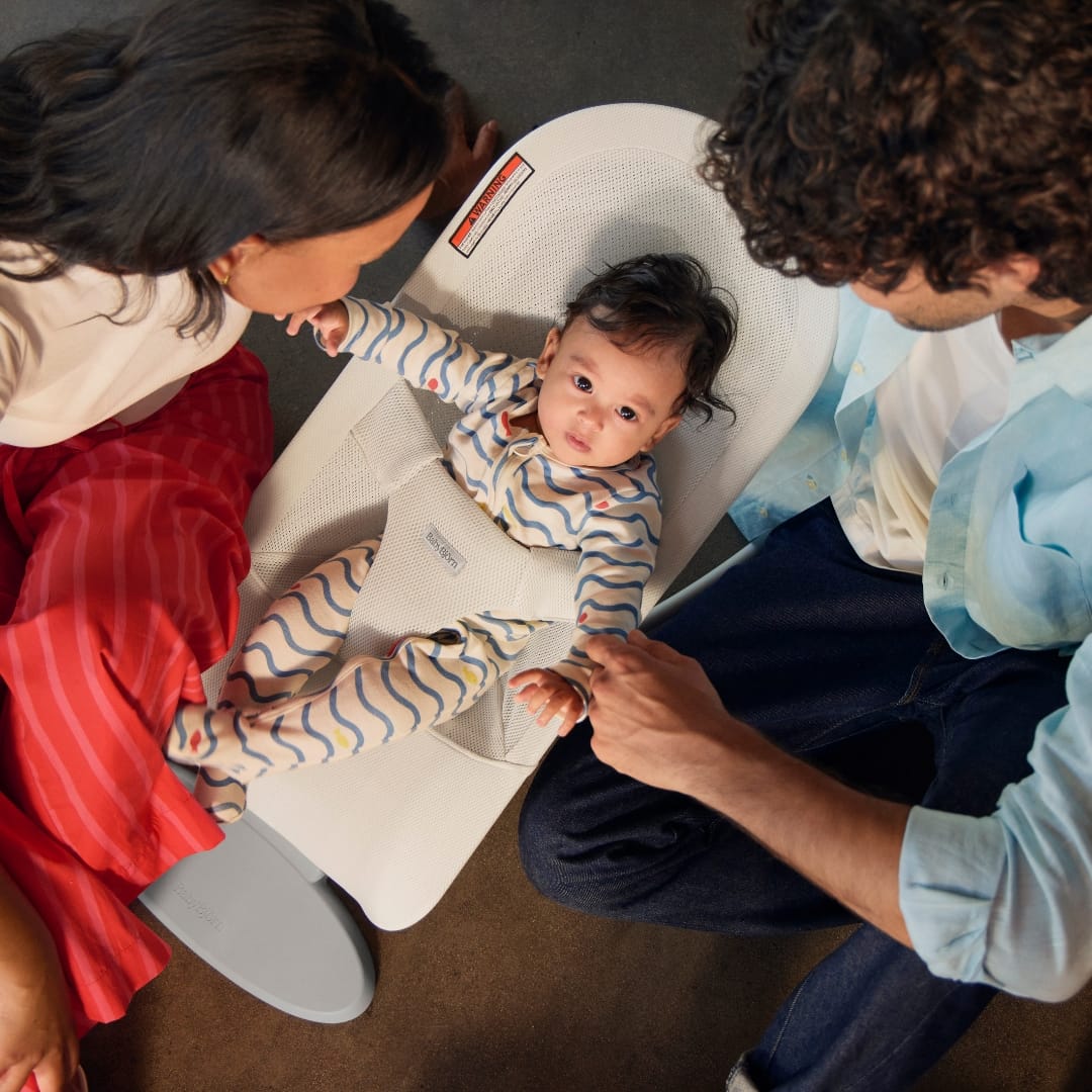 Baby in a beige BabyBjorn bouncer with parents sitting on the floor