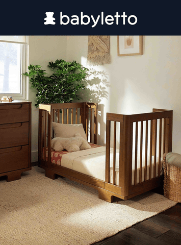 A brown wooden babyletto crib sits on a beige rug in a warmly lit room. "babyletto" is printed at the top.