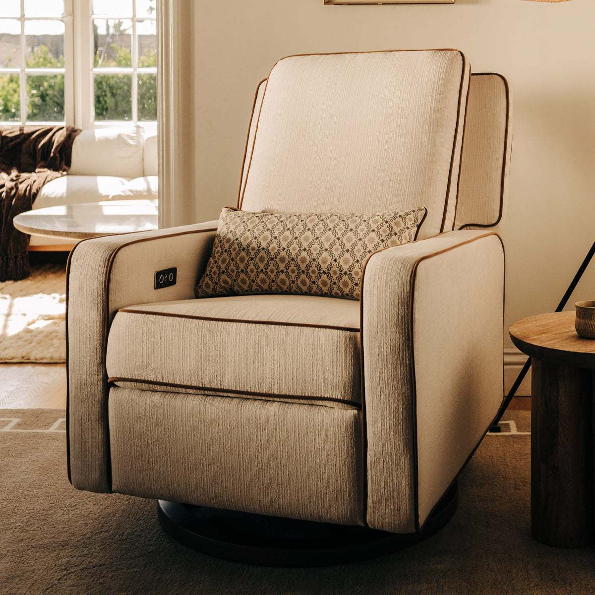 Babyletto beige swivel glider recliner with patterned lumbar pillow and brown piping