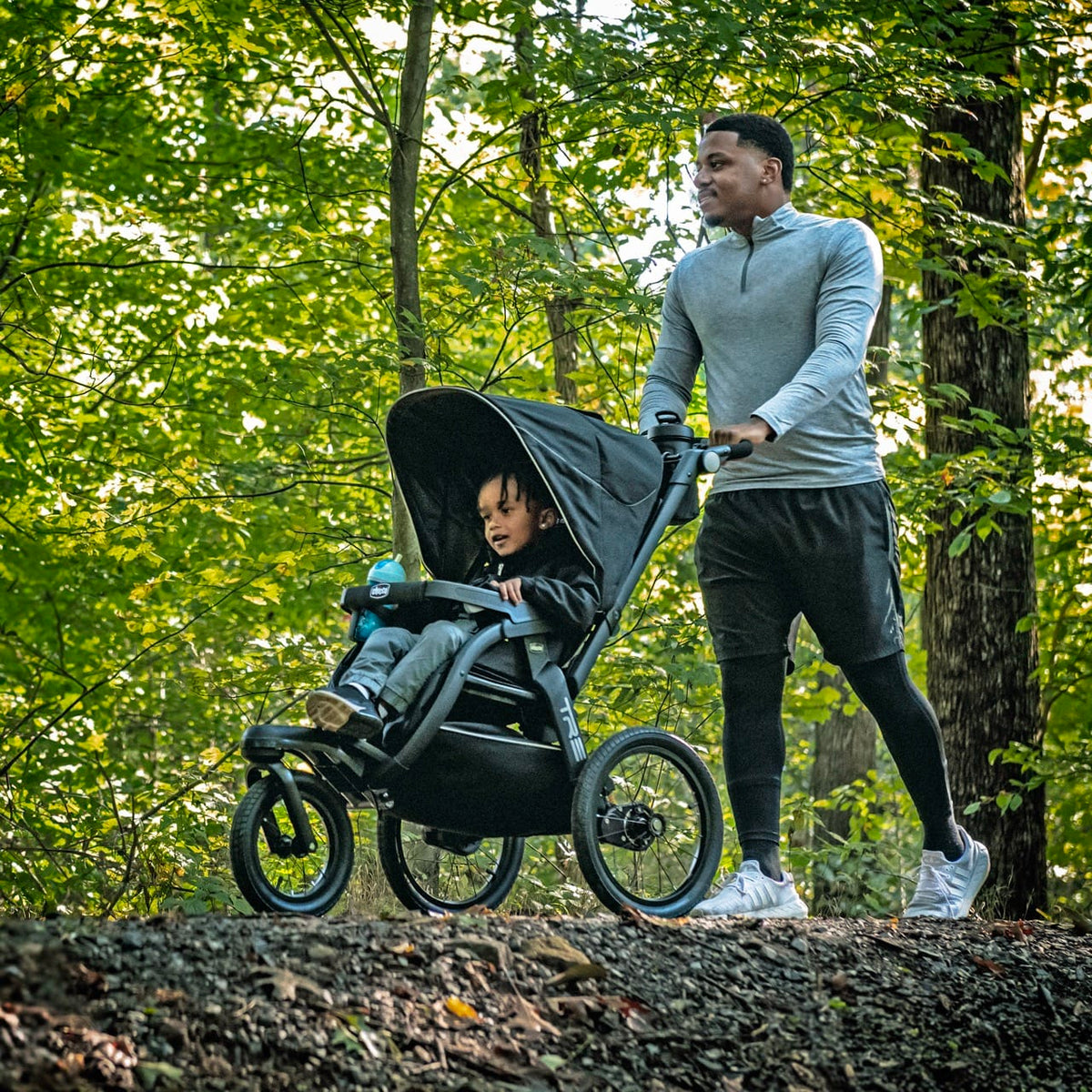 Man pushing a child in a black Chicco jogging stroller on a wooded trail