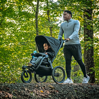 Man pushing a child in a black Chicco jogging stroller on a wooded trail