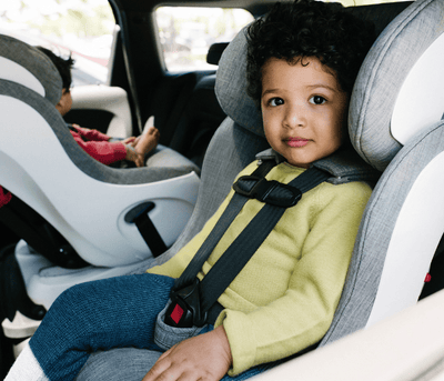 A young child with curly hair sits buckled in a gray car seat, wearing a yellow shirt and blue jeans. Another child is visible in the background.
