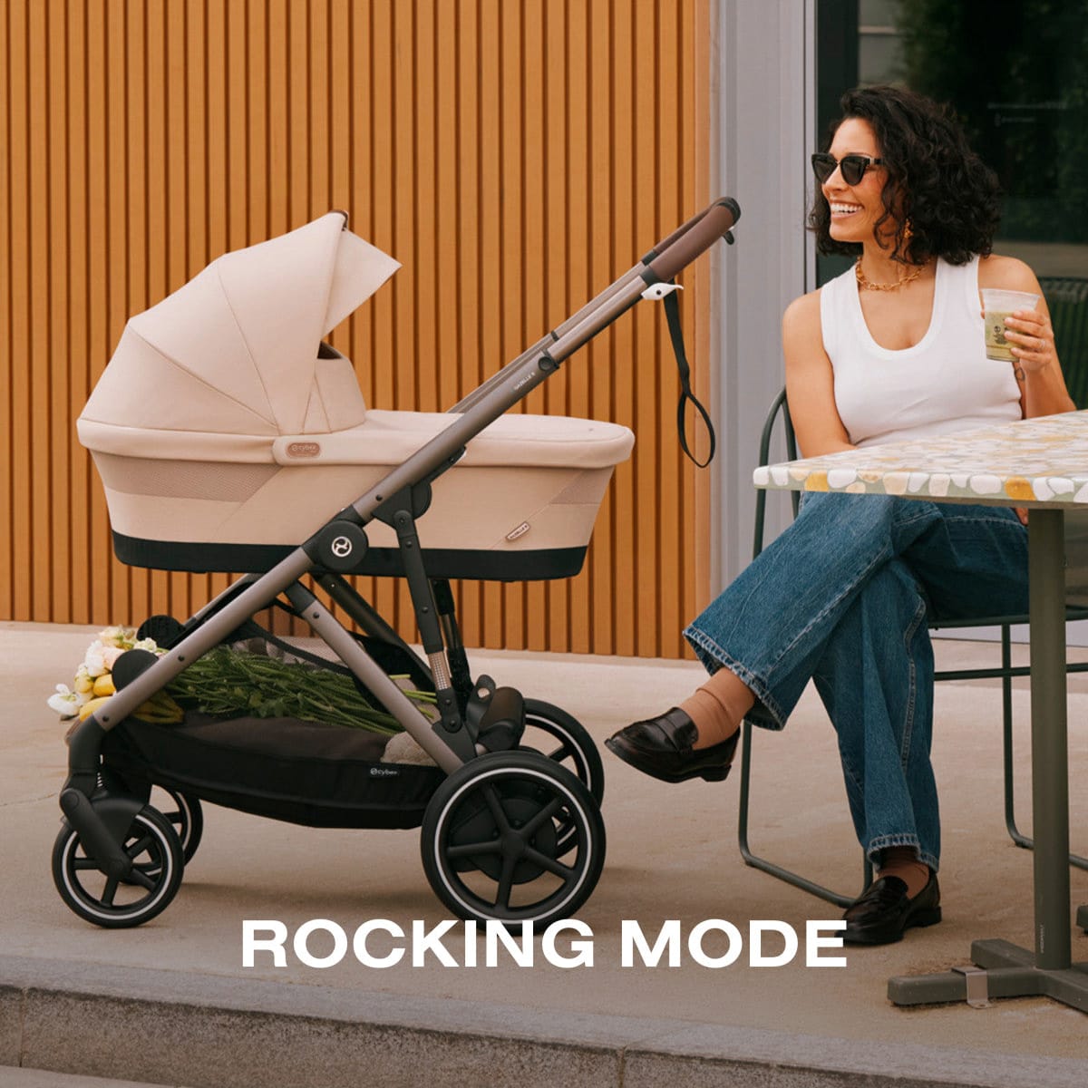 Cybex Gazelle S stroller with beige carrycot in rocking mode next to a woman sitting at a cafe table