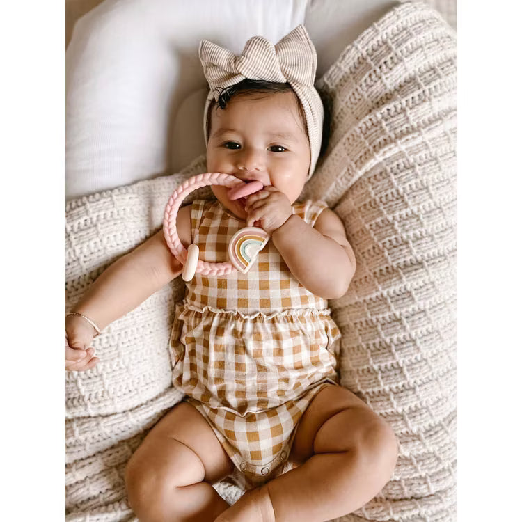 Baby wearing a bow headband and gingham romper holding an Itzy Ritzy rainbow silicone teether