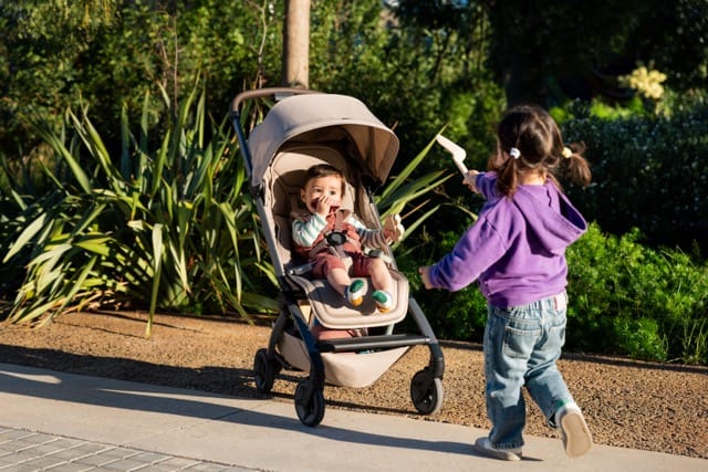 Child in a Joolz stroller with a sibling playing nearby in a park