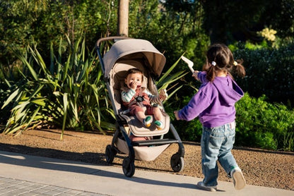 Child in a Joolz stroller with a sibling playing nearby in a park
