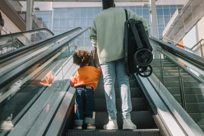 Person carrying a folded Joolz stroller on an escalator with a child