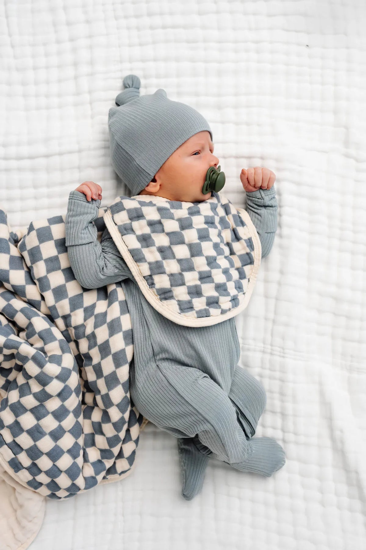 Newborn wearing a blue ribbed sleeper and hat with a checkered bib and matching blanket
