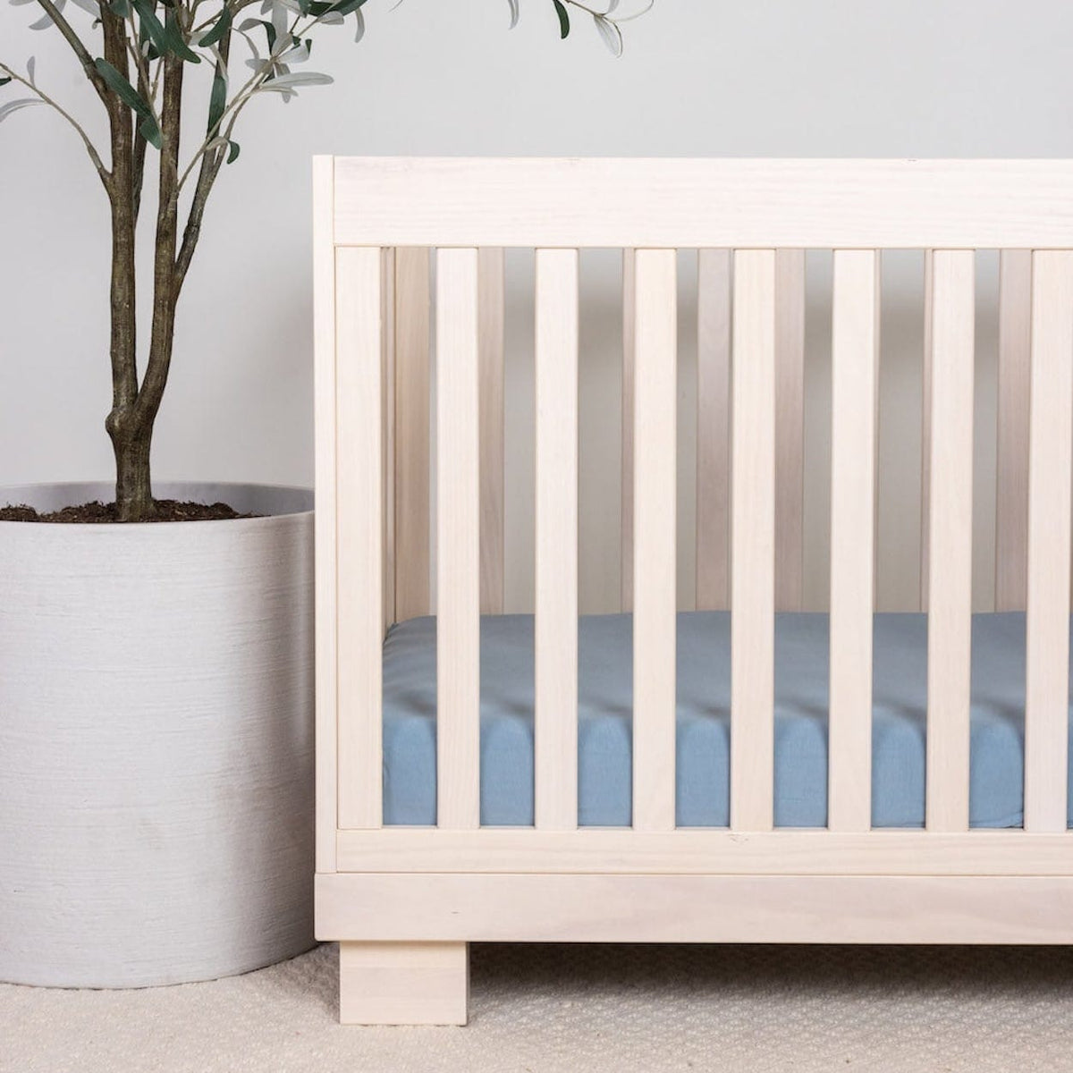 Light wood crib with blue bedding next to a potted plant