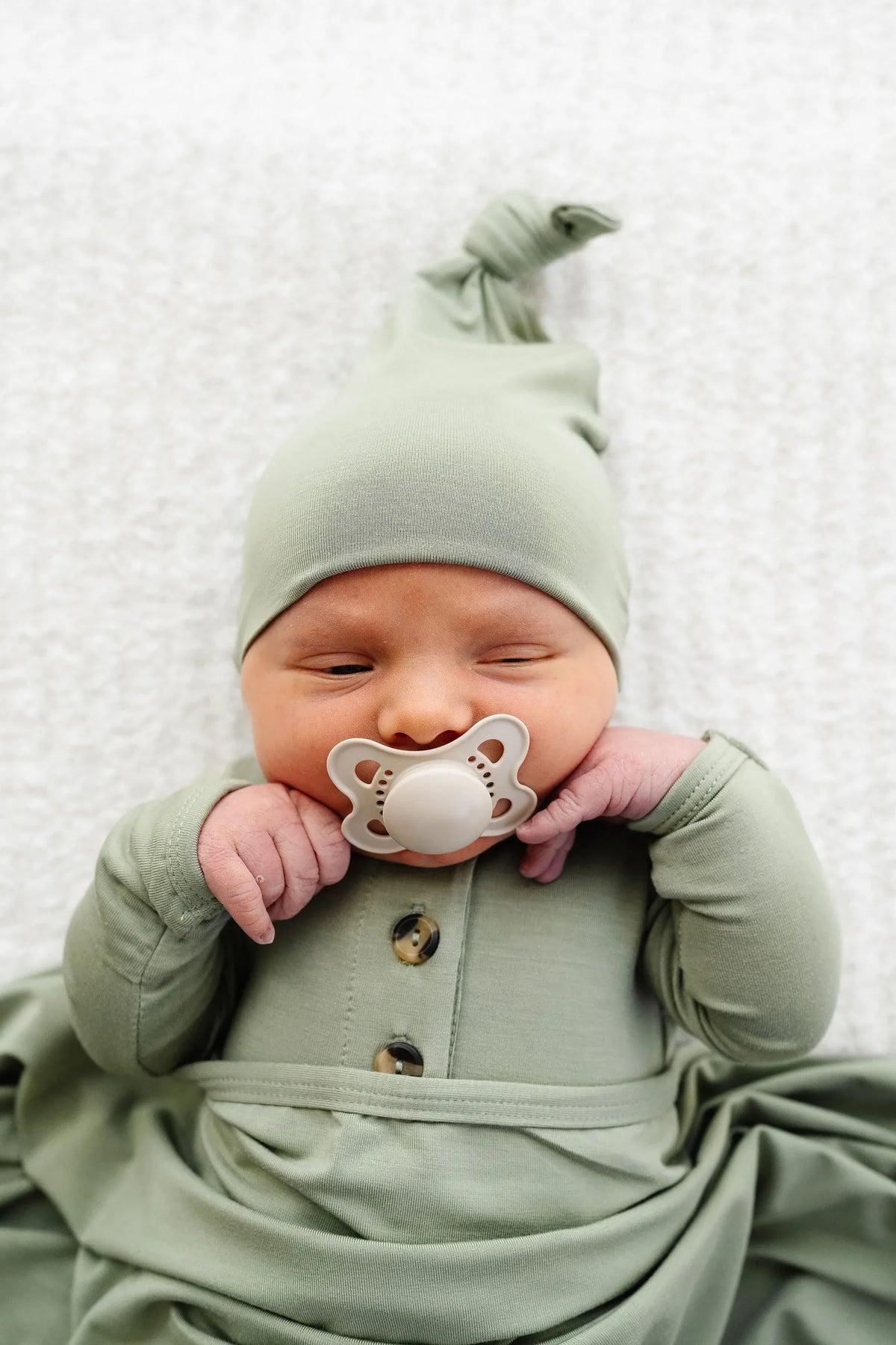 Newborn wearing a sage green knotted hat and matching buttoned sleeper with a pacifier