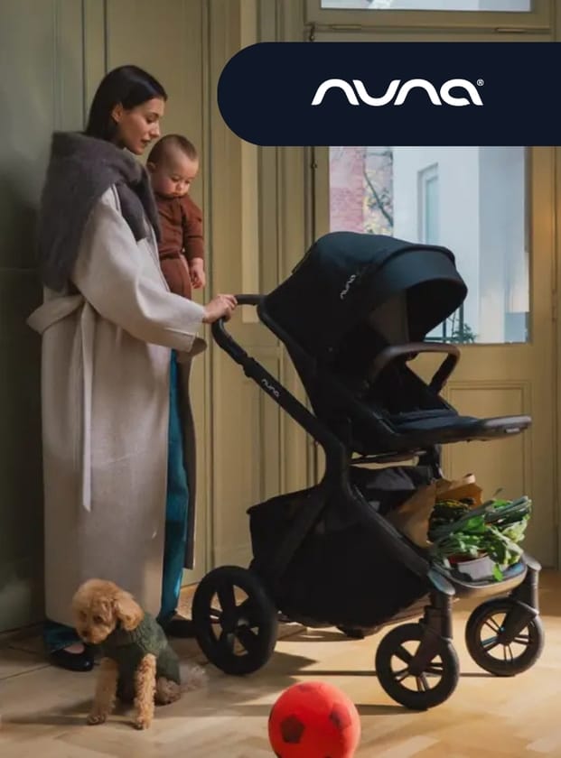 Woman in beige coat holds baby beside black Nuna stroller with groceries, small dog nearby, red ball on floor, warm indoor lighting.
