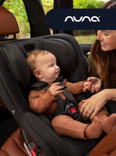 A baby in a black car seat looks up at a woman, who is smiling at the baby. Visible text: "nuna".